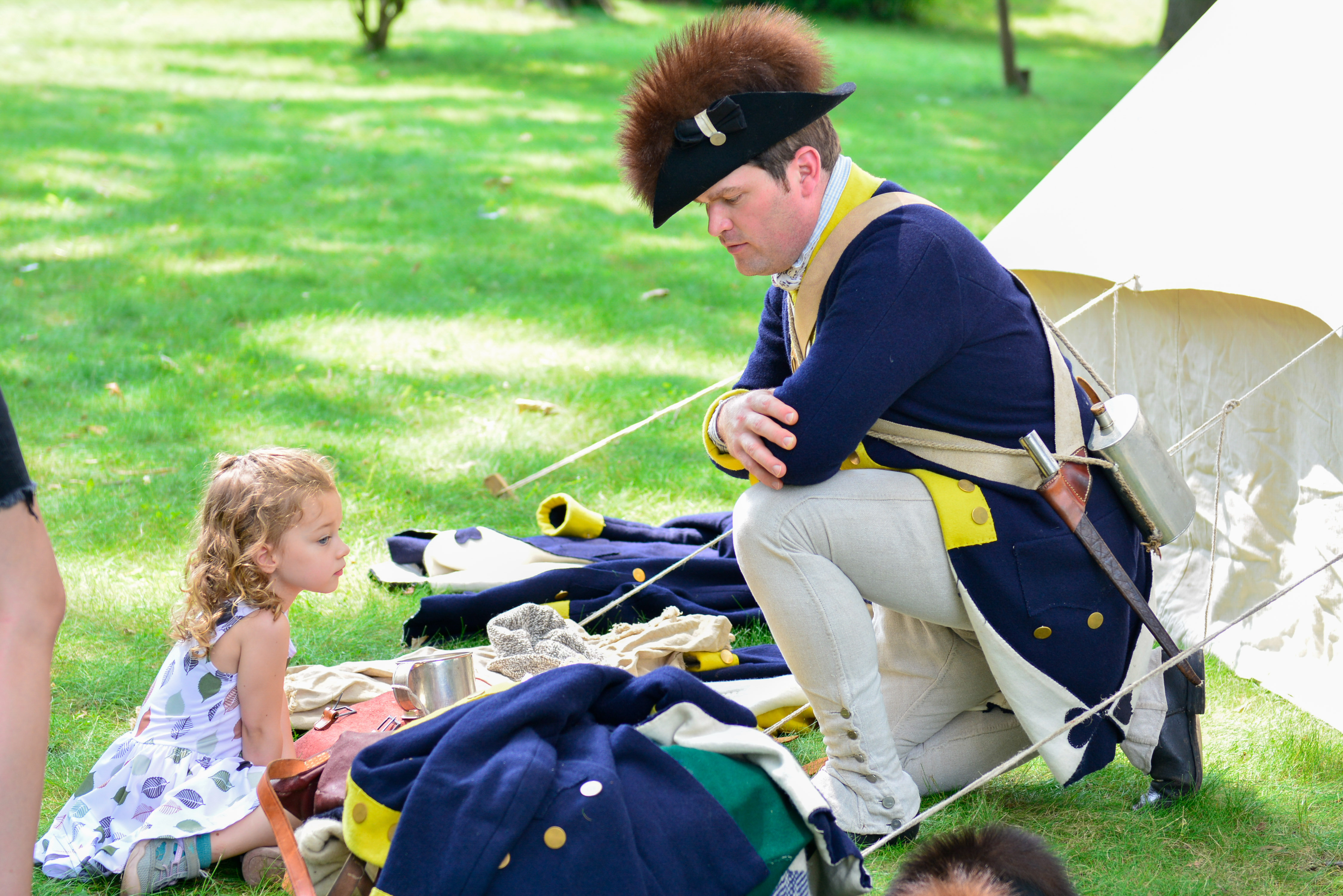 Costumed living history interpreter Joel Anderson kneels to the right of the image wearing a blue and gold commander in chief's guard jacket and blacket and fur lined hat while talking to a young visitor at Morristown National Historical Park.