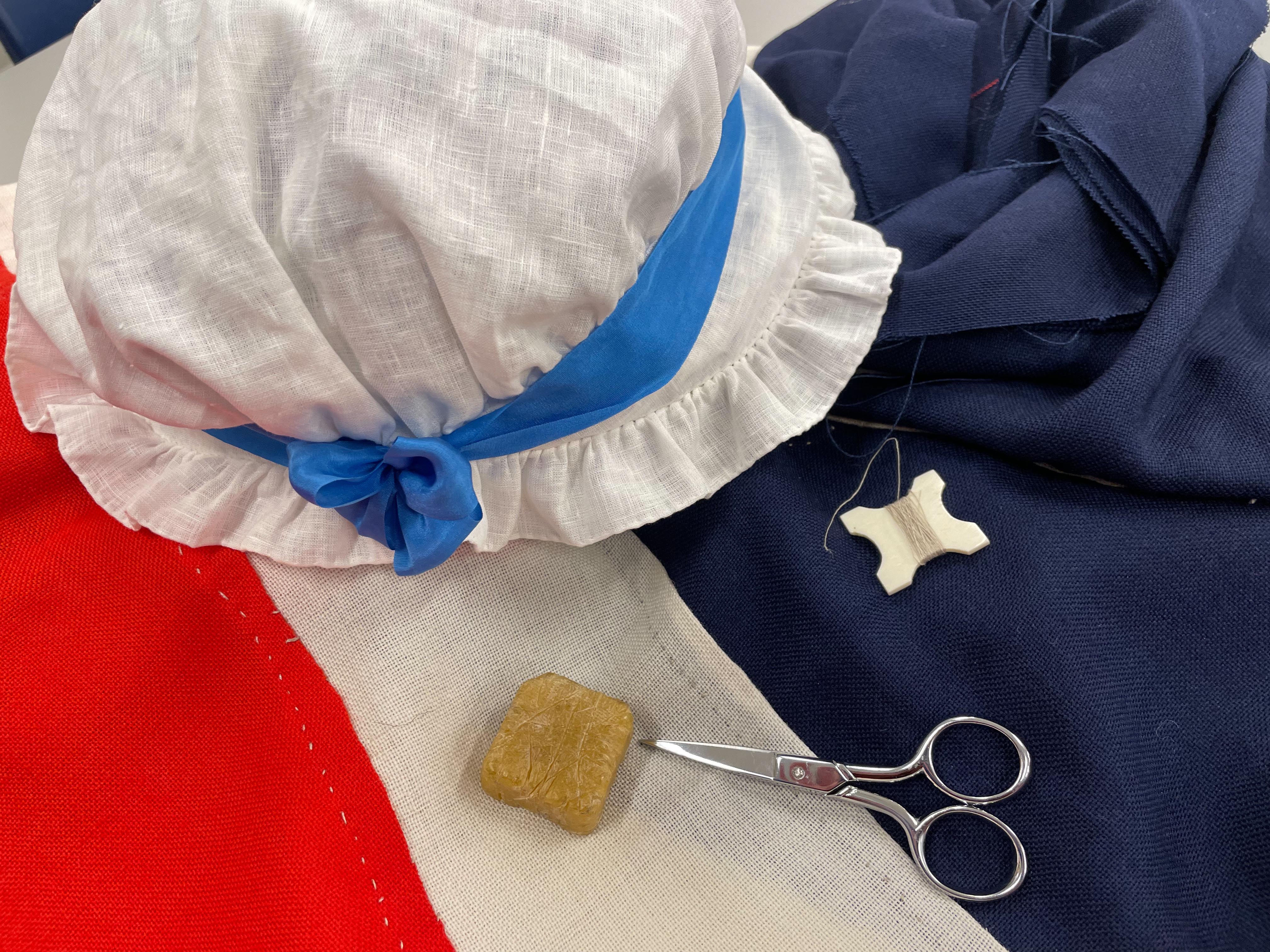 image of red, which, and blue fabric, a white bonnet, scissors, thread, and other flag making tools.