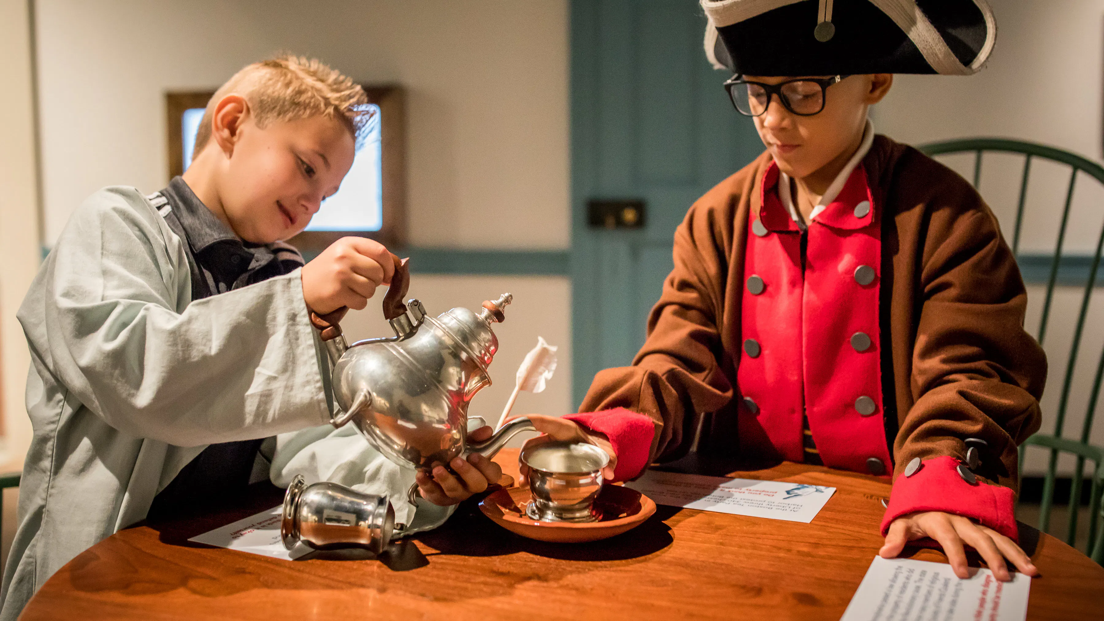 Two white male adolescents, both standing at a wooden table, interact with a play tea set. The adolescent on the left is pretending to pour tea into a silver teacup from a silver teapot. The adolescent on the right has his hand on the teacup saucer. Both are dressed in Revolutionary-era outfits.