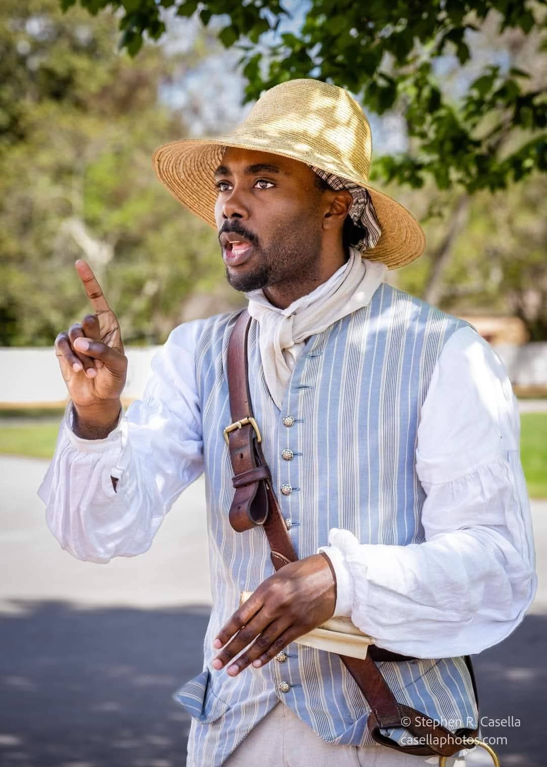A Black man in a white shirt, blue-and-white striped vest, and straw hat pointing while speaking