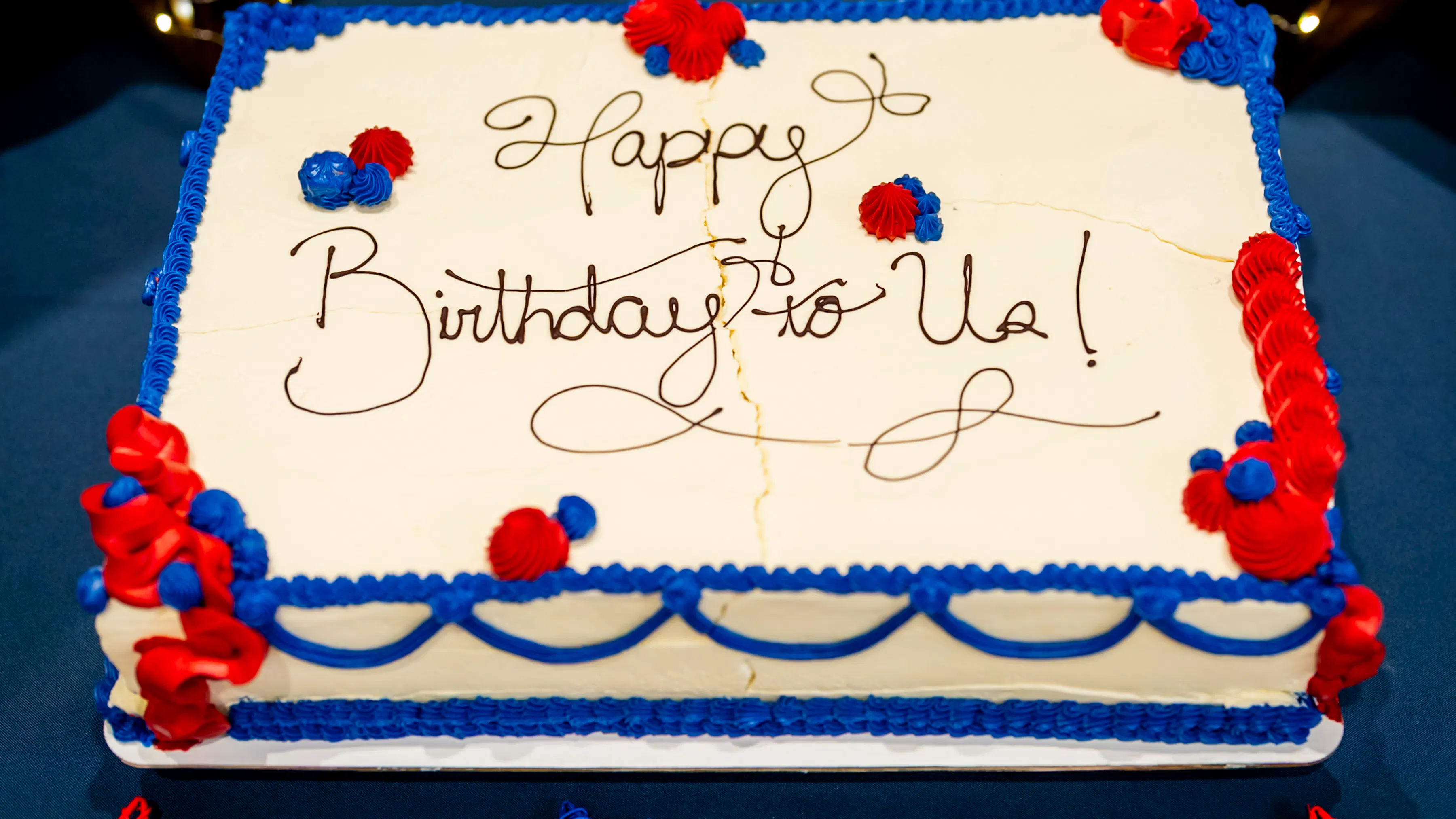 An image of a white sheet cake with red and blue frosting decorations and "Happy Birthday to Us!" written in icing on the top