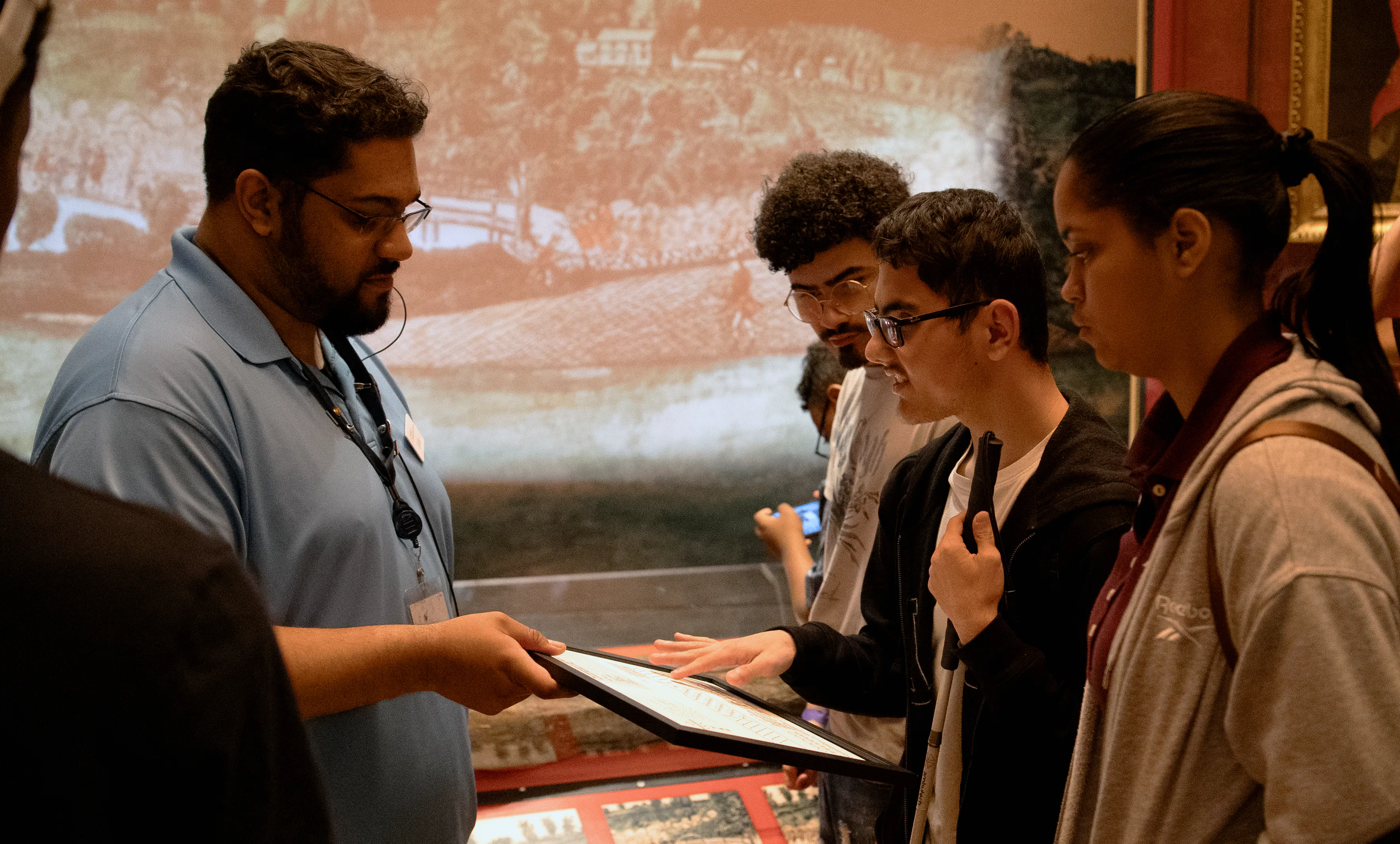 A man in a blue shirt holds a white board in a black frame for a man holding a cane