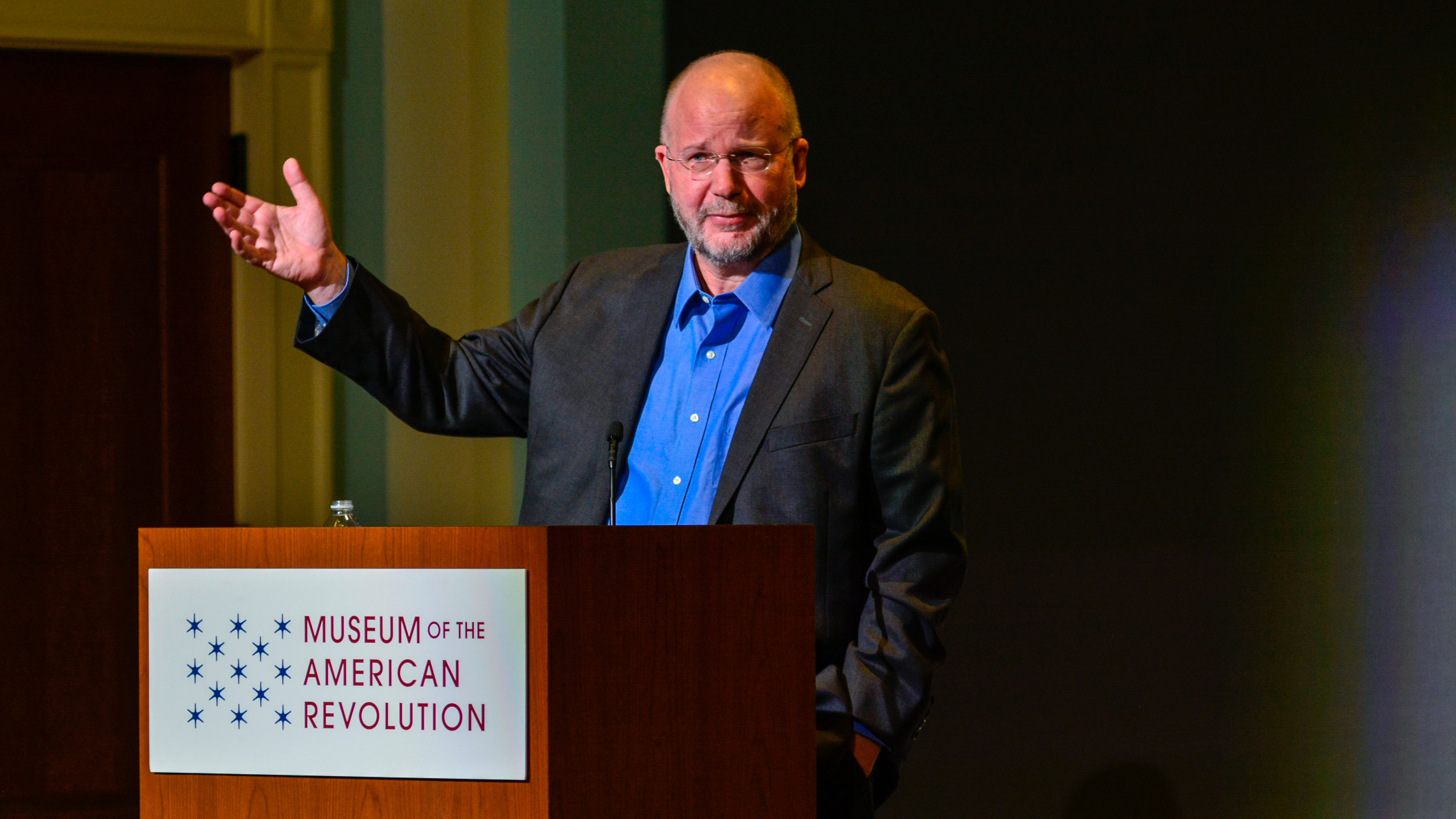William Hogeland, a white man with glasses and a salt and pepper beard, speaks from a podium with the Museum logo on it.