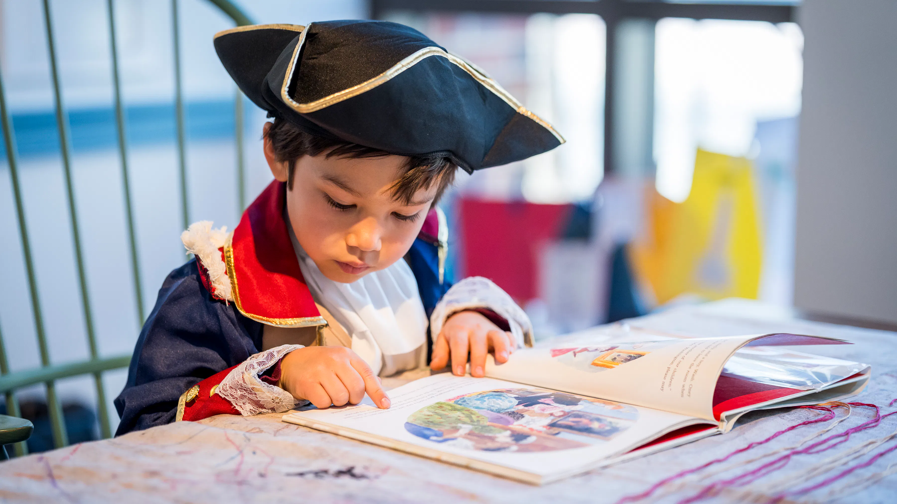 Child wearing a tricorn hat and red and navy blue coat reads a children's book