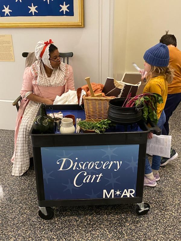 Costumed living historian Leslie Bramlett sitting in a chair at left of the image wearing 18th century pink and white dress talking to a young visitor at a museum discovery cart with replica objects.
