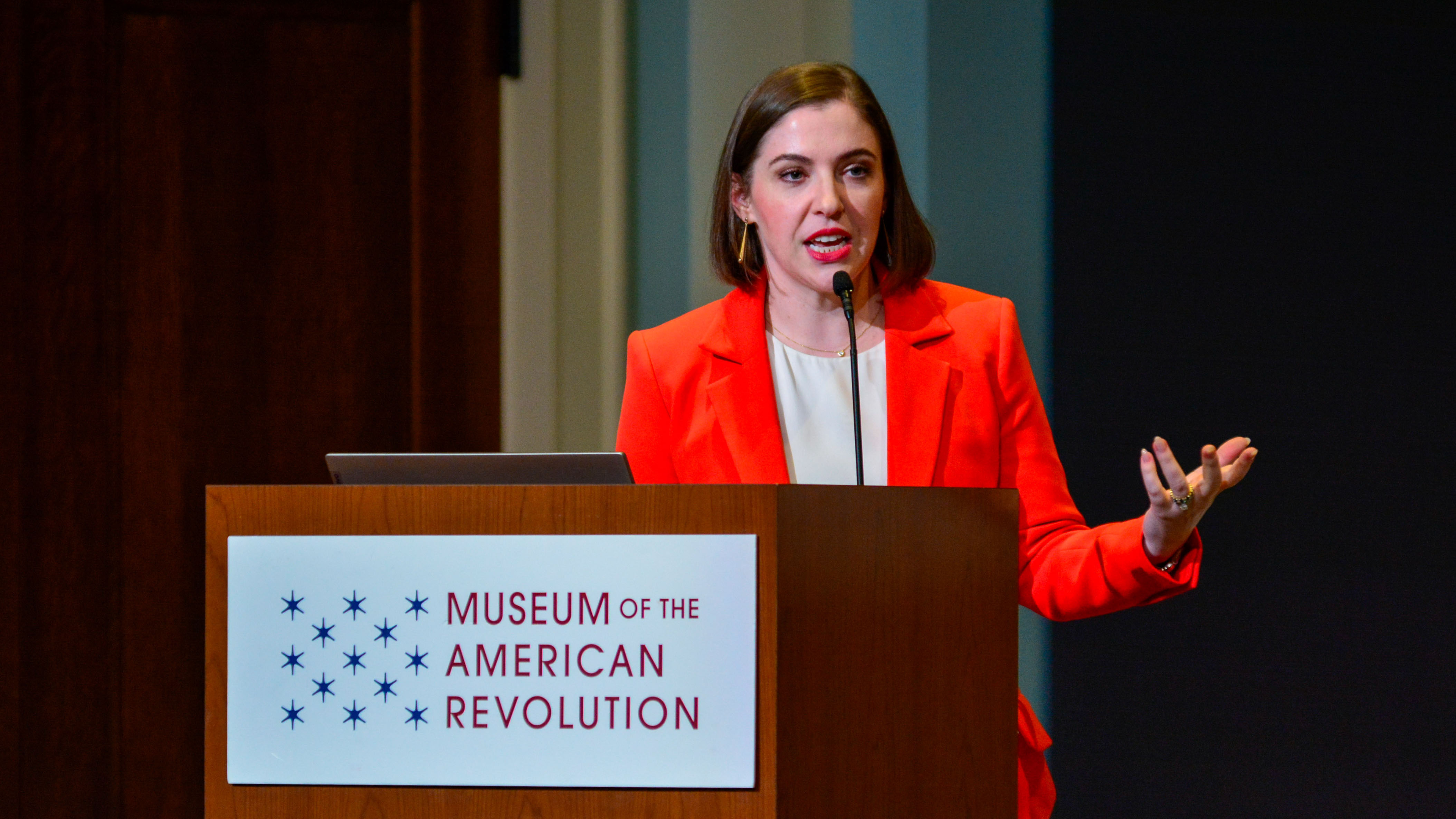 This image shows Dr. Lindsay M. Chervinsky standing at a wooden podium with the Museum's logo on the front while wearing a red suit.