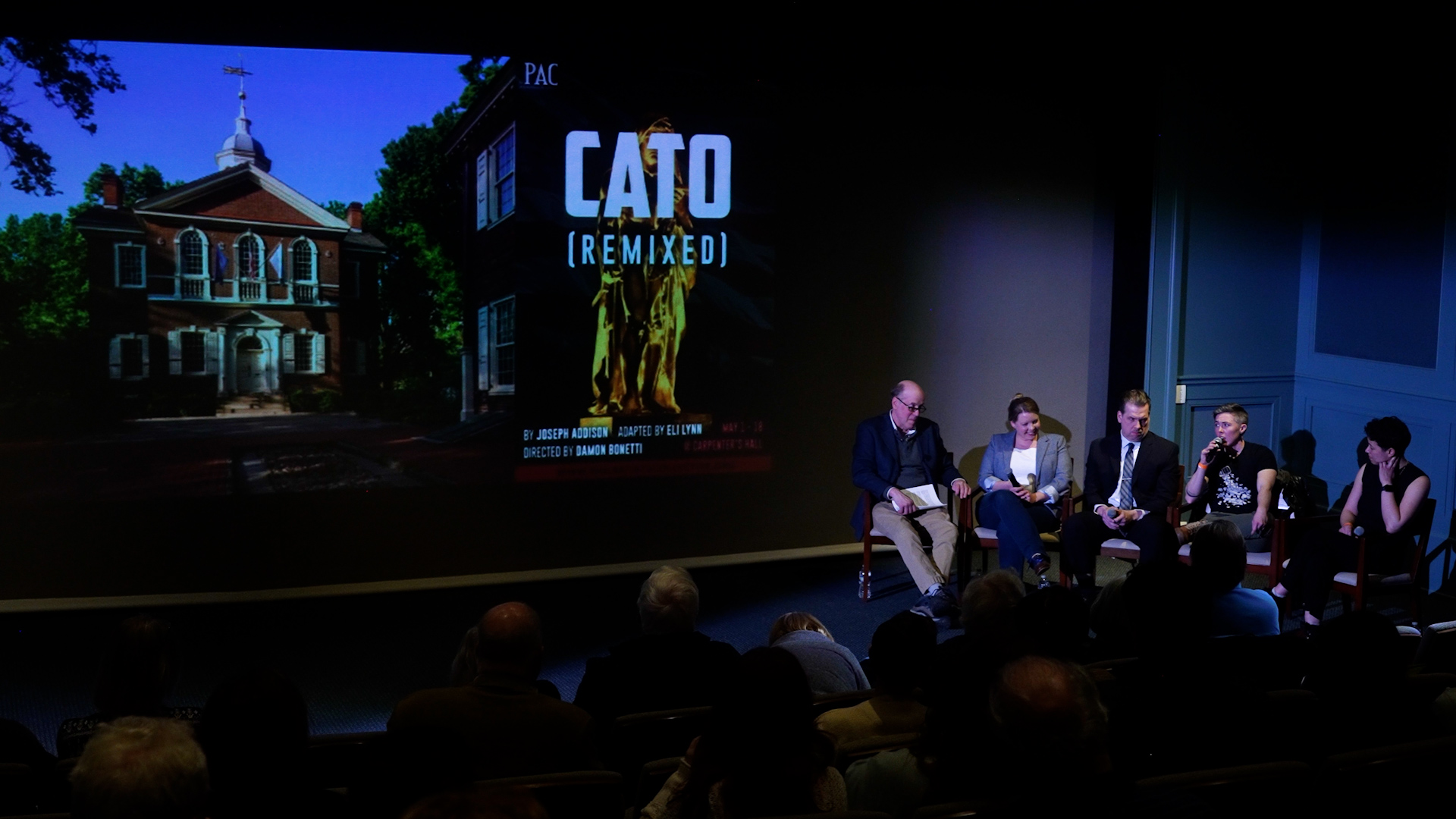 A photo of five people in chairs at the right. There is an image of Carpenters' Hall alongside the words CATO: REMIXED projected on a screen on the back wall.