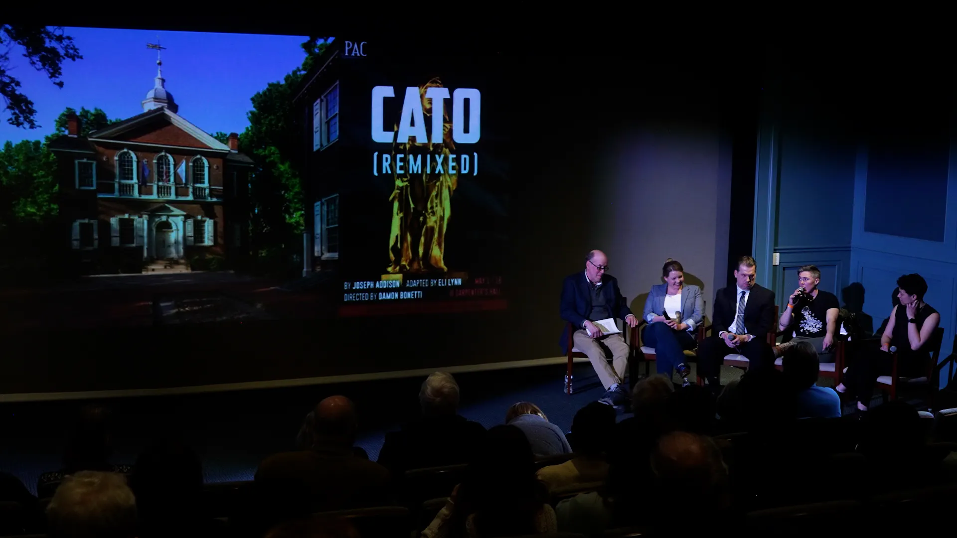 A photo of five people in chairs at the right. There is an image of Carpenters' Hall alongside the words CATO: REMIXED projected on a screen on the back wall.