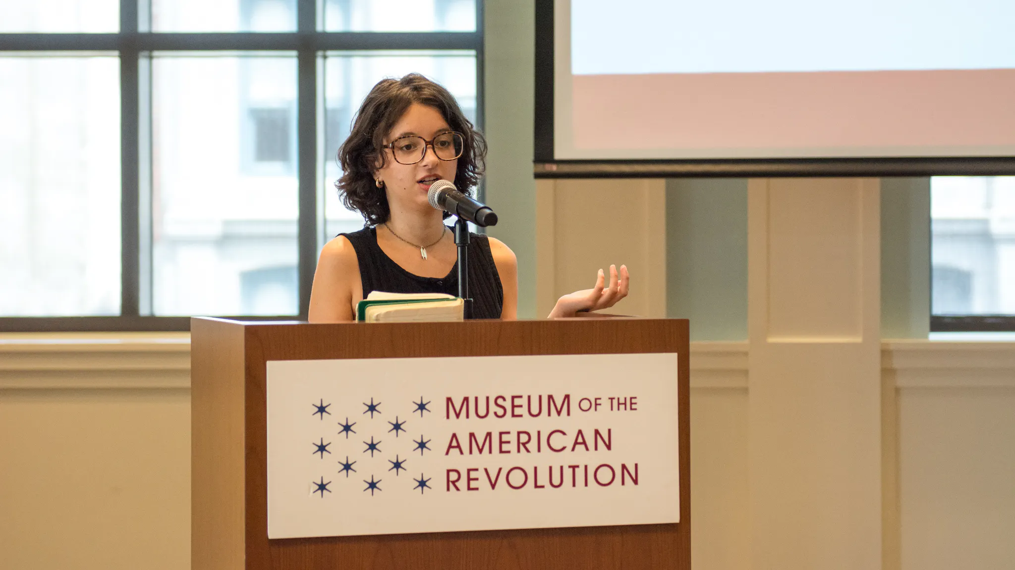 A young white woman with dark hair and glasses speaks at a podium