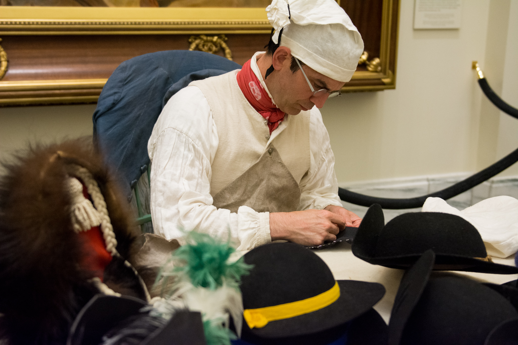 A man in a white 18th-century shirt and cap sewing hats while seated at a table