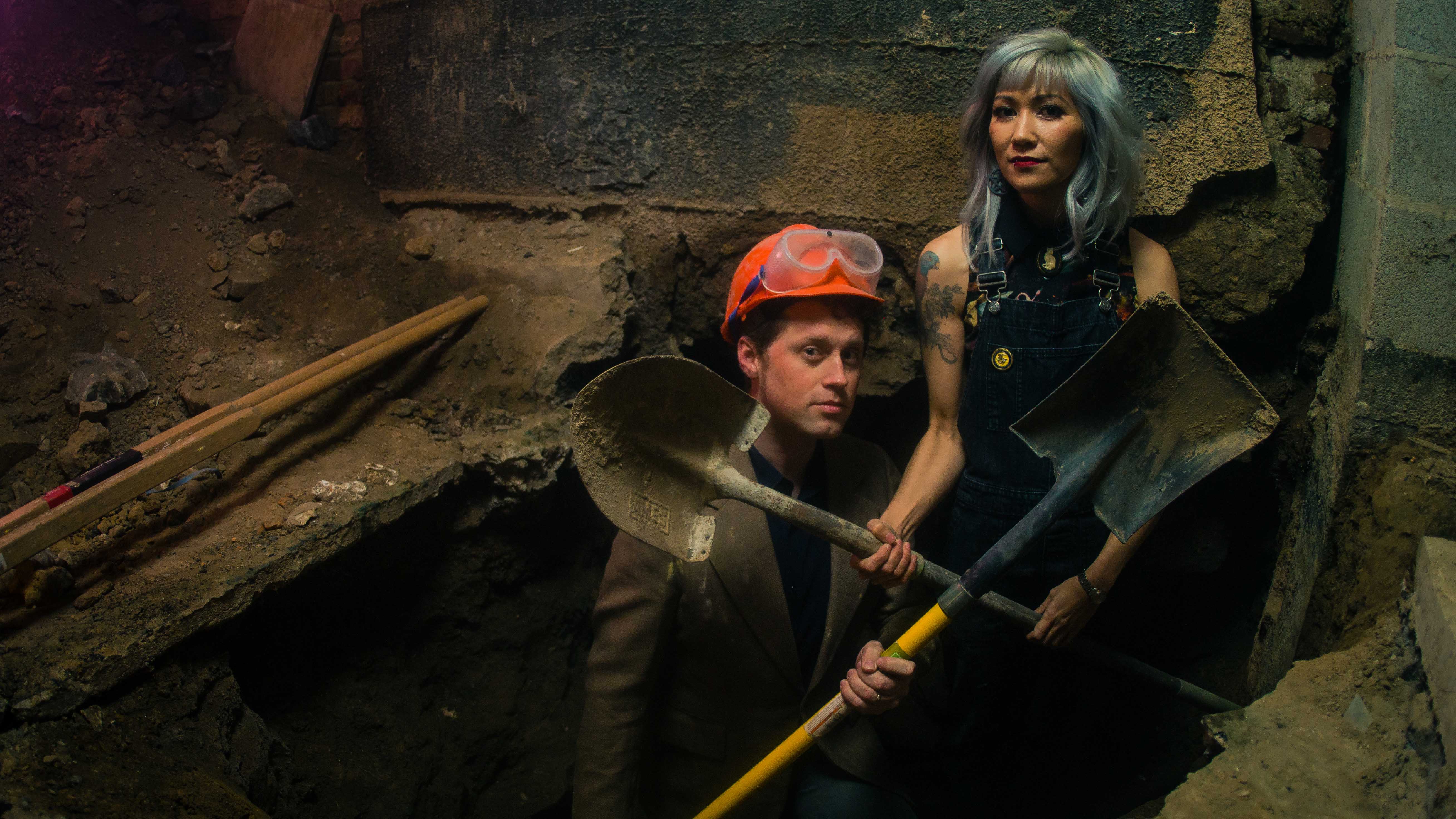 Matt and Melissa Dunphy holding shovels and standing in a hole they dug in the basement of the theater they purchased.