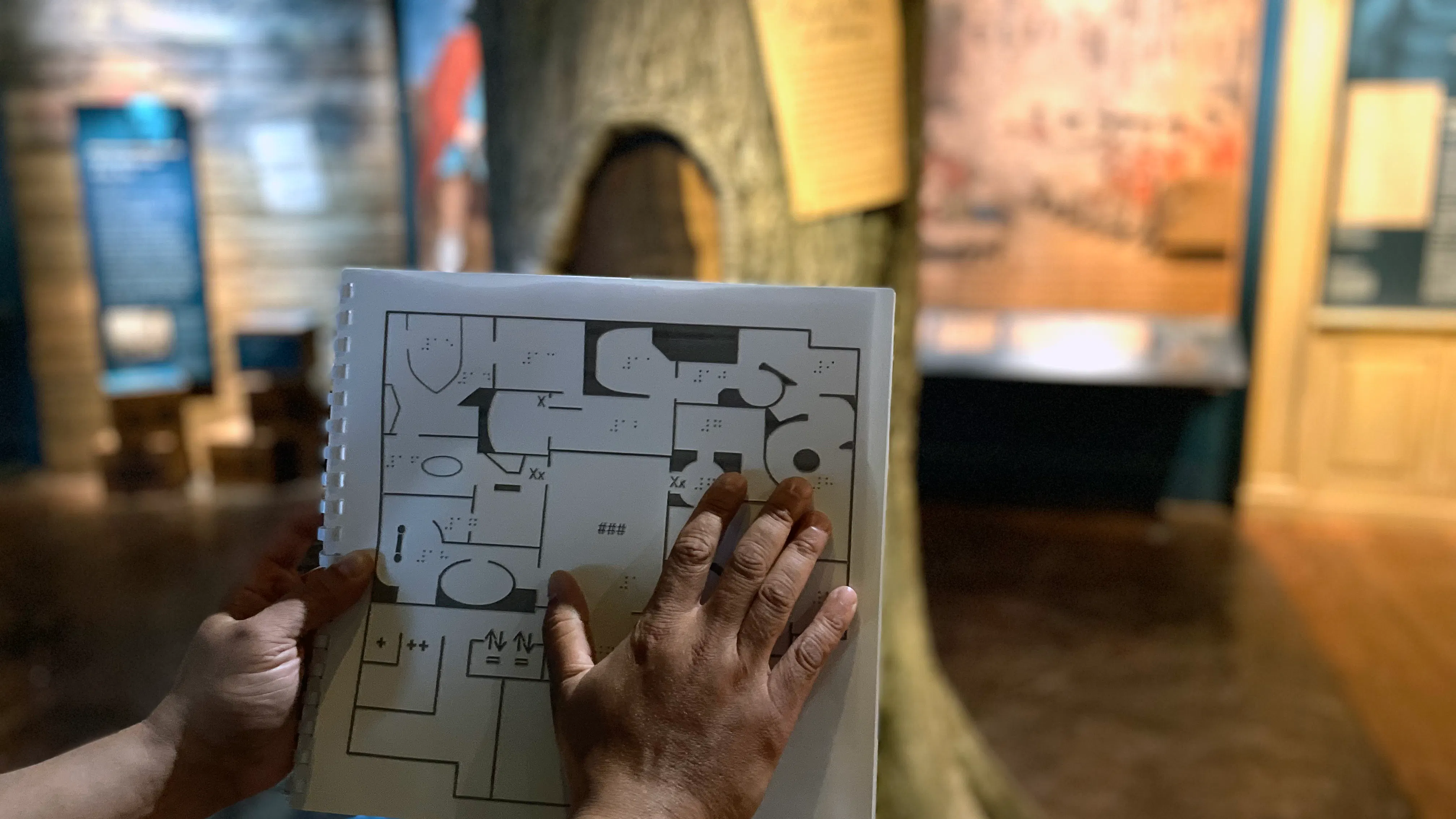 A hand holding a map of the Museum's core galleries including Braille descriptions with a Liberty Tree in the background.
