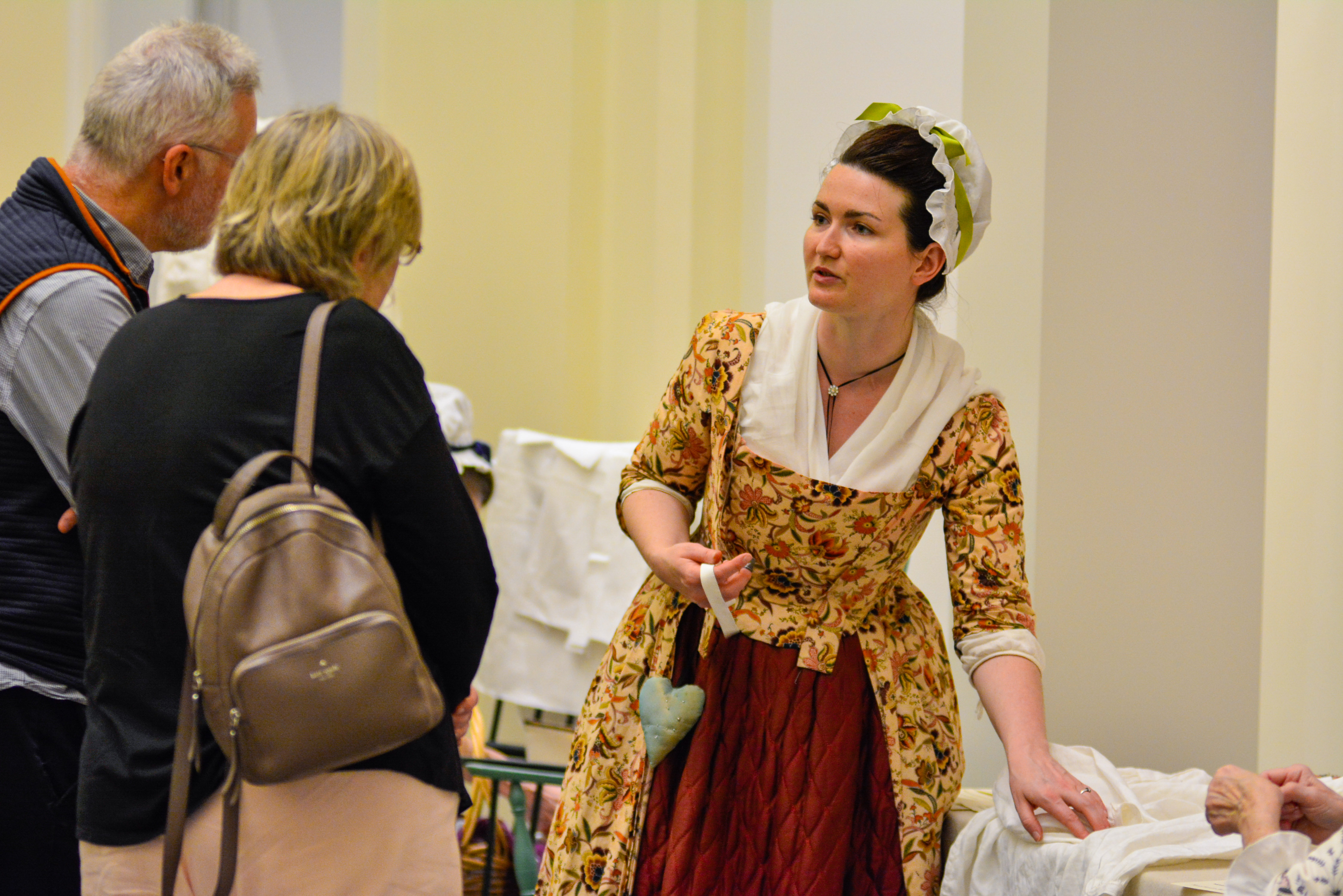 A costumed historical interpreter talks to two guests about sewing at a table in the Museum rotunda.