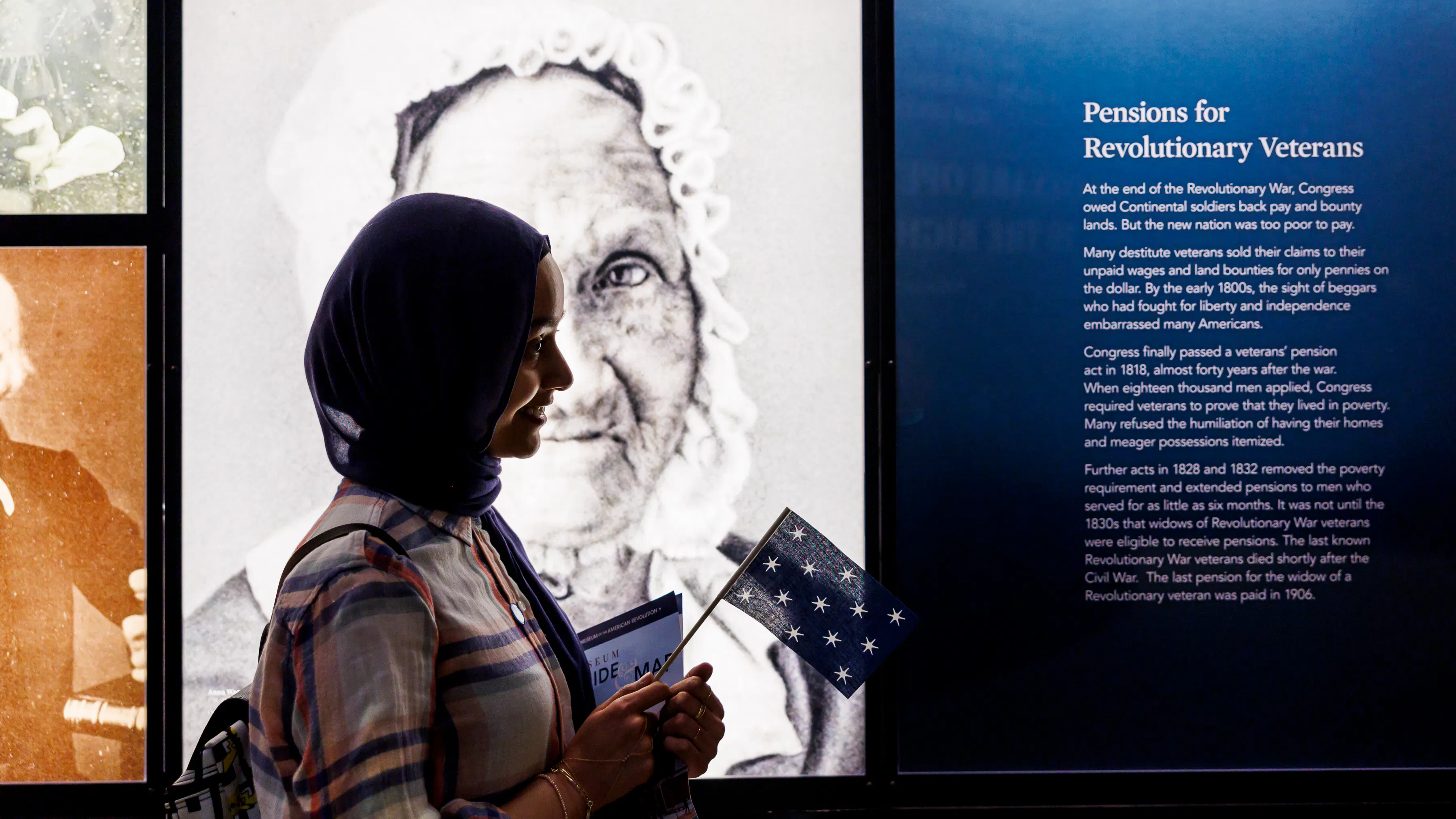 A visitor holds a miniature version of George Washington's standard flag in front of photographs of people from the Revolutionary generation who lived into the age of photography.