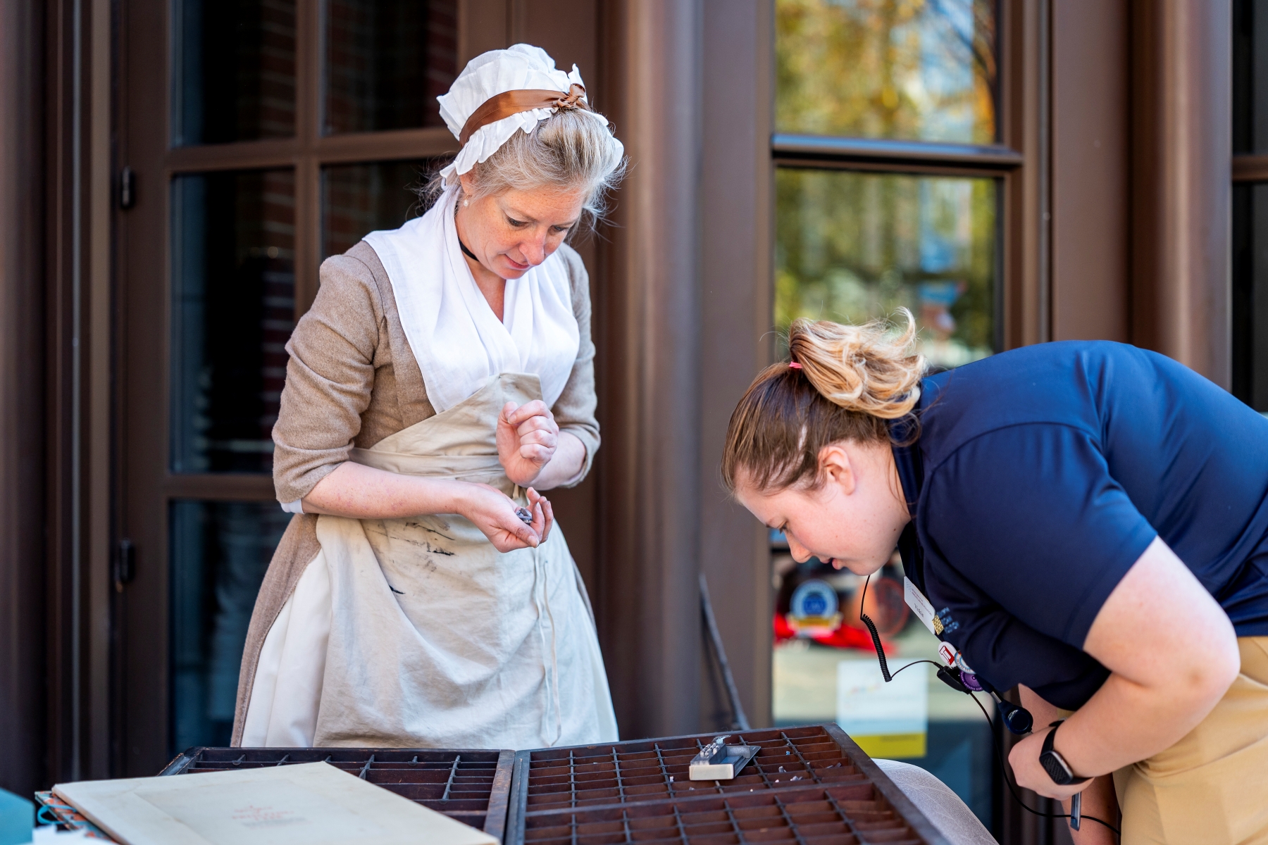 A woman in 18th century dress stands behind a table of typesetting implements while a staff member looks at them