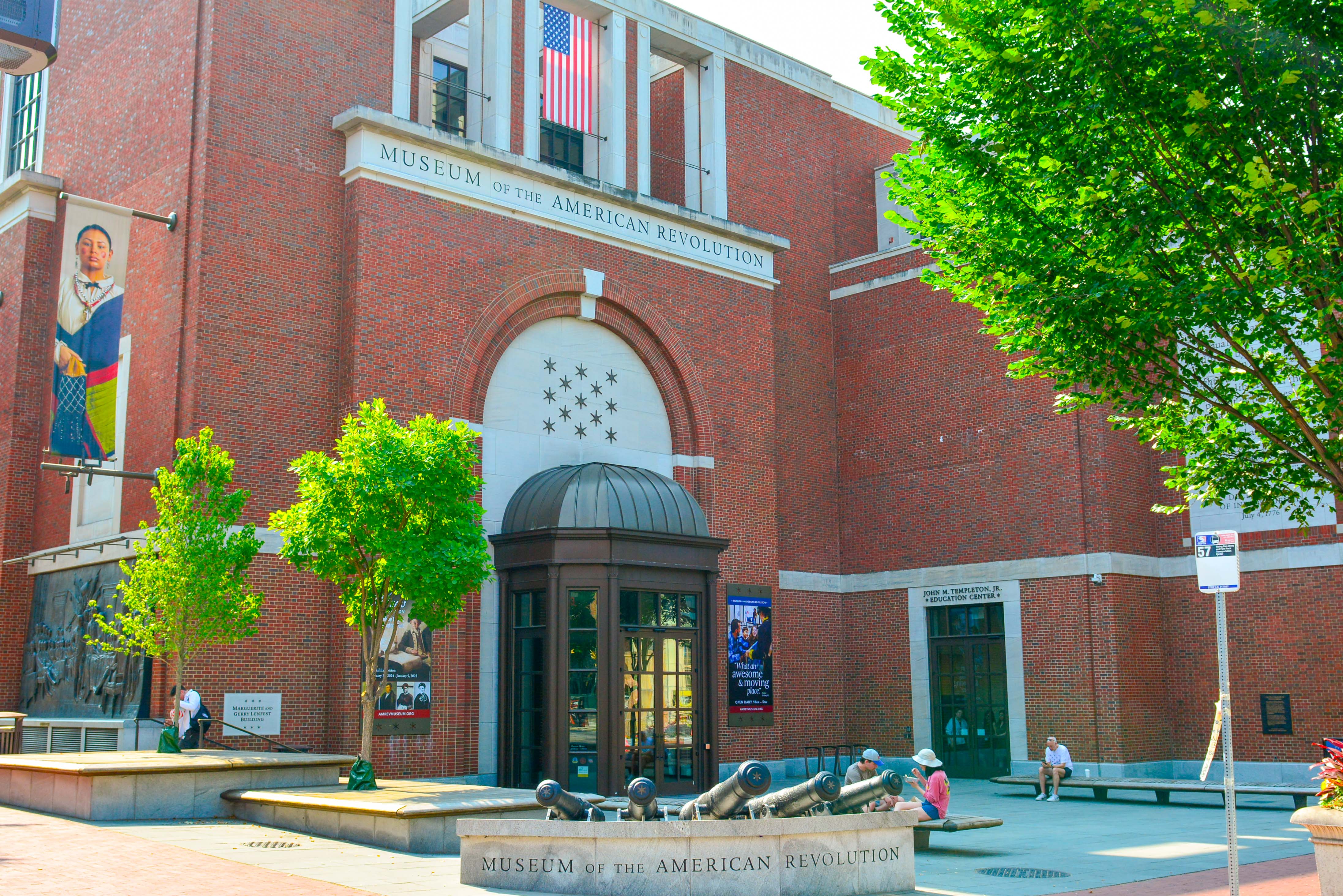 This image shows the red brick exterior of the Museum of the American Revolution with green trees on either side of the entrance.