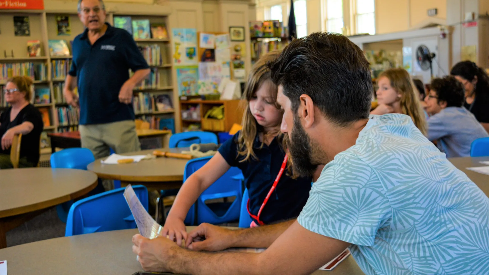 An older man and young child read while a man standing lectures in the background.