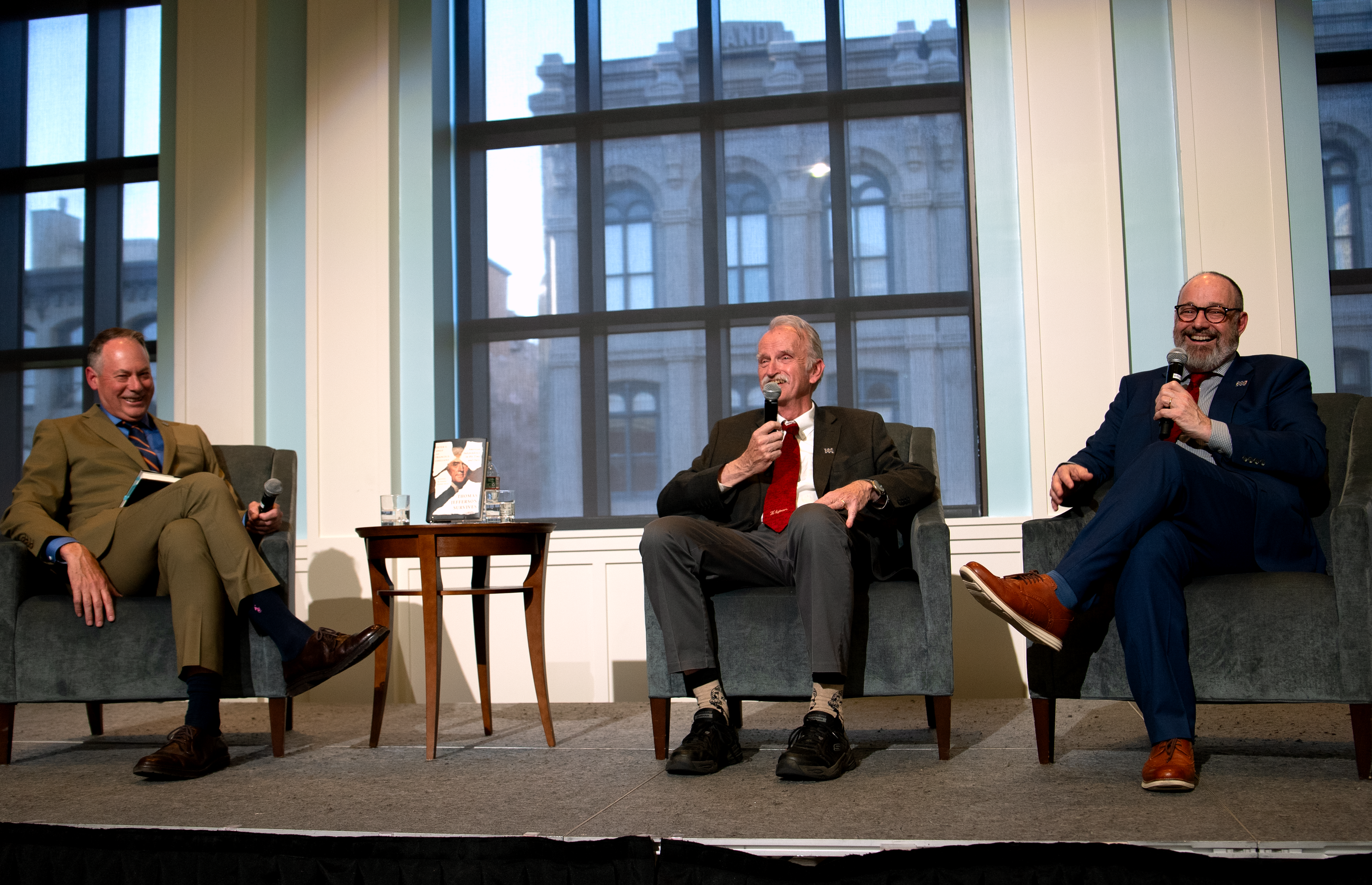 Three white men sit on stage in front of large windows