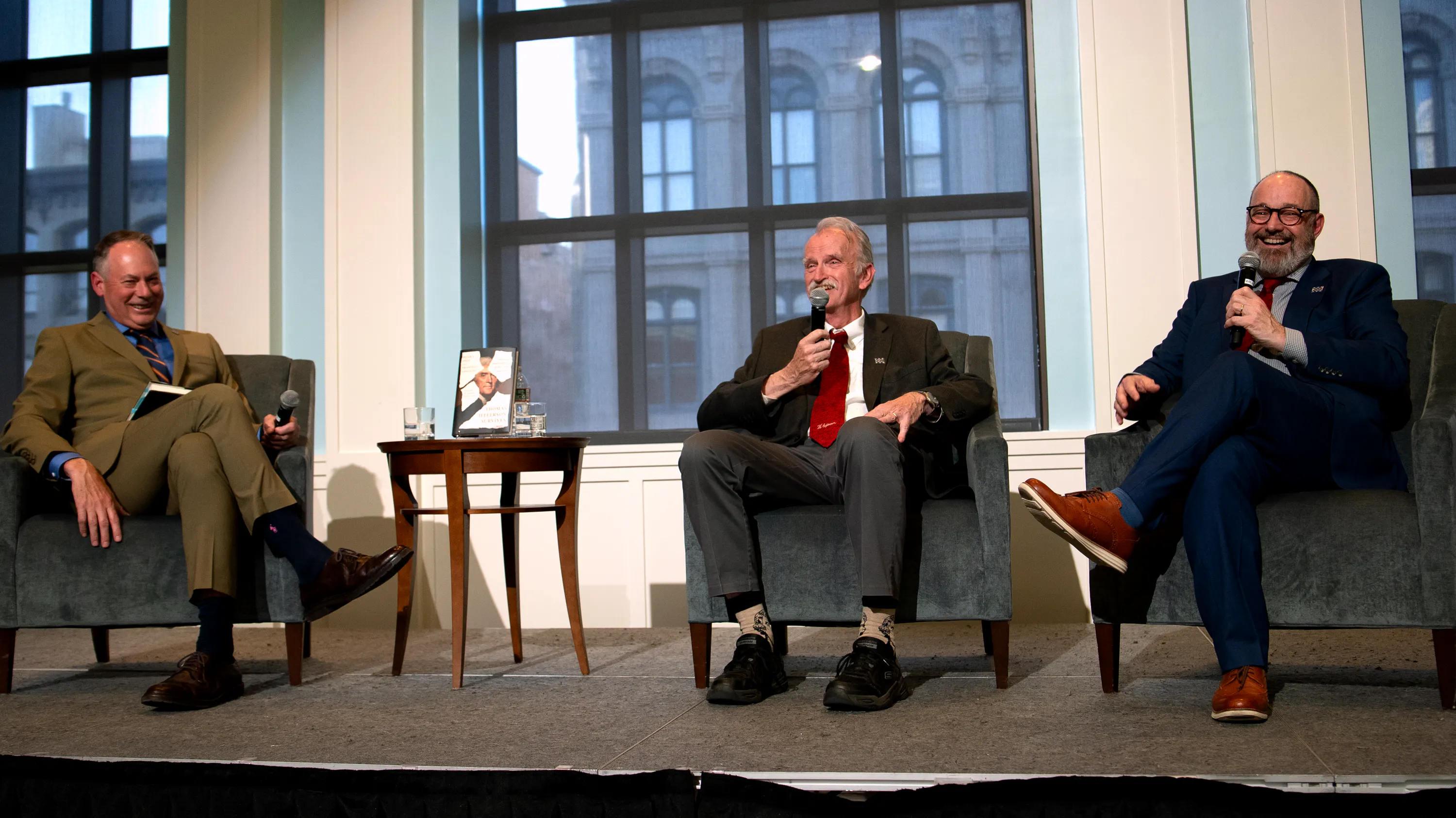 Three white men sit on stage in front of large windows
