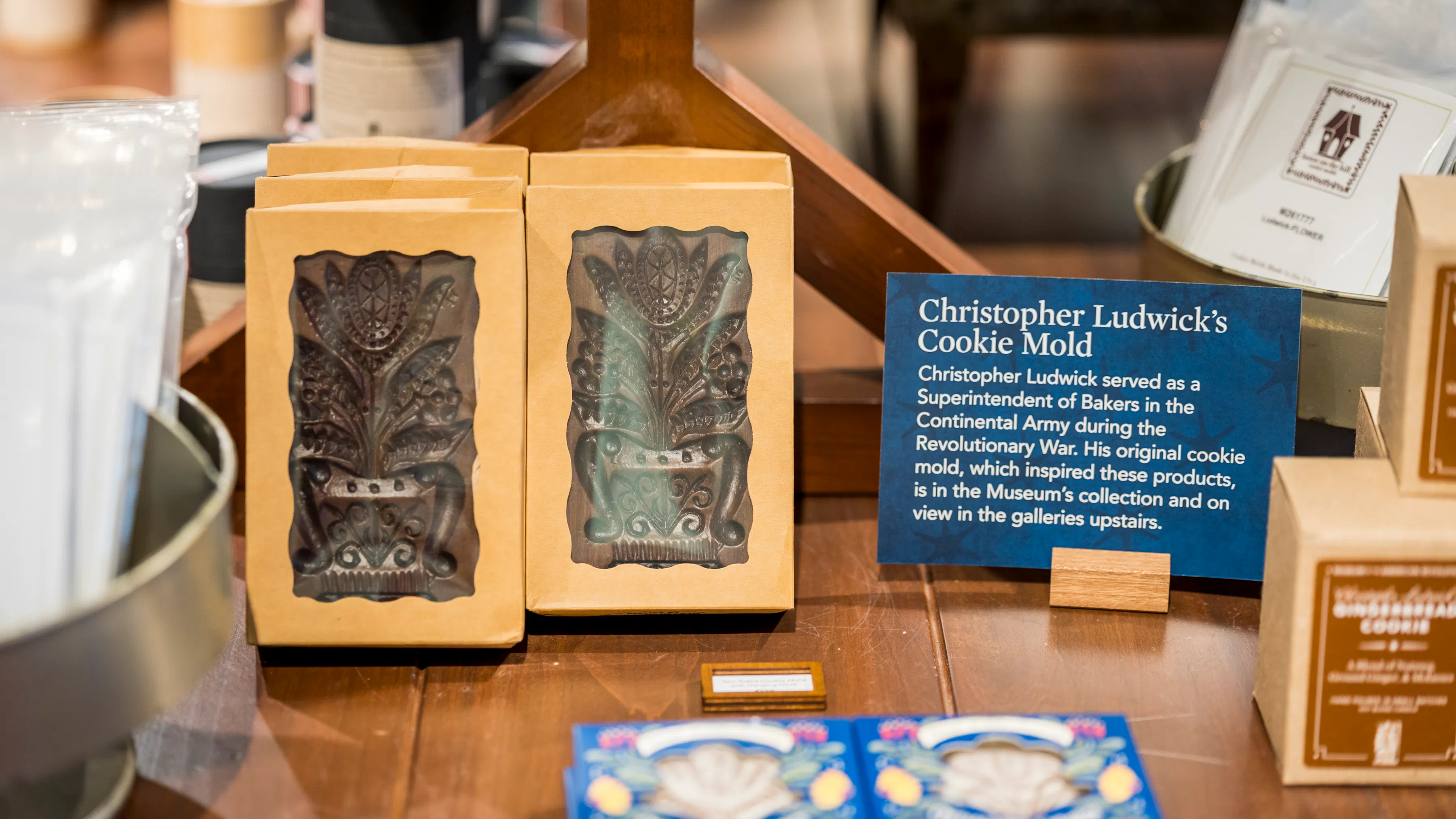 A blue sign reads "Christopher Ludwick's Cookie Mold" next to a display of brown cookie molds in tan boxes.