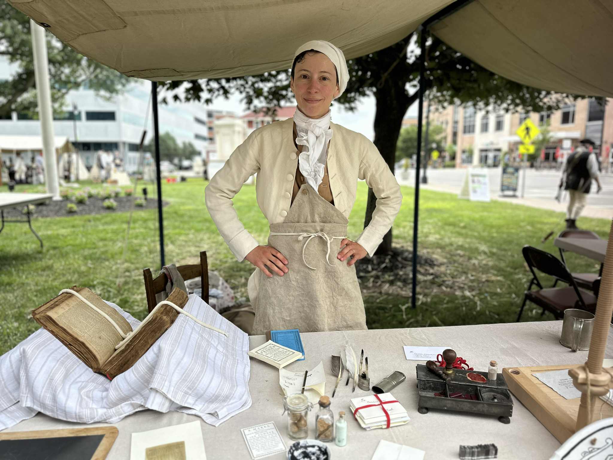A costumed interpreter stands in an outdoor tent behind a table with bookmaking supplies