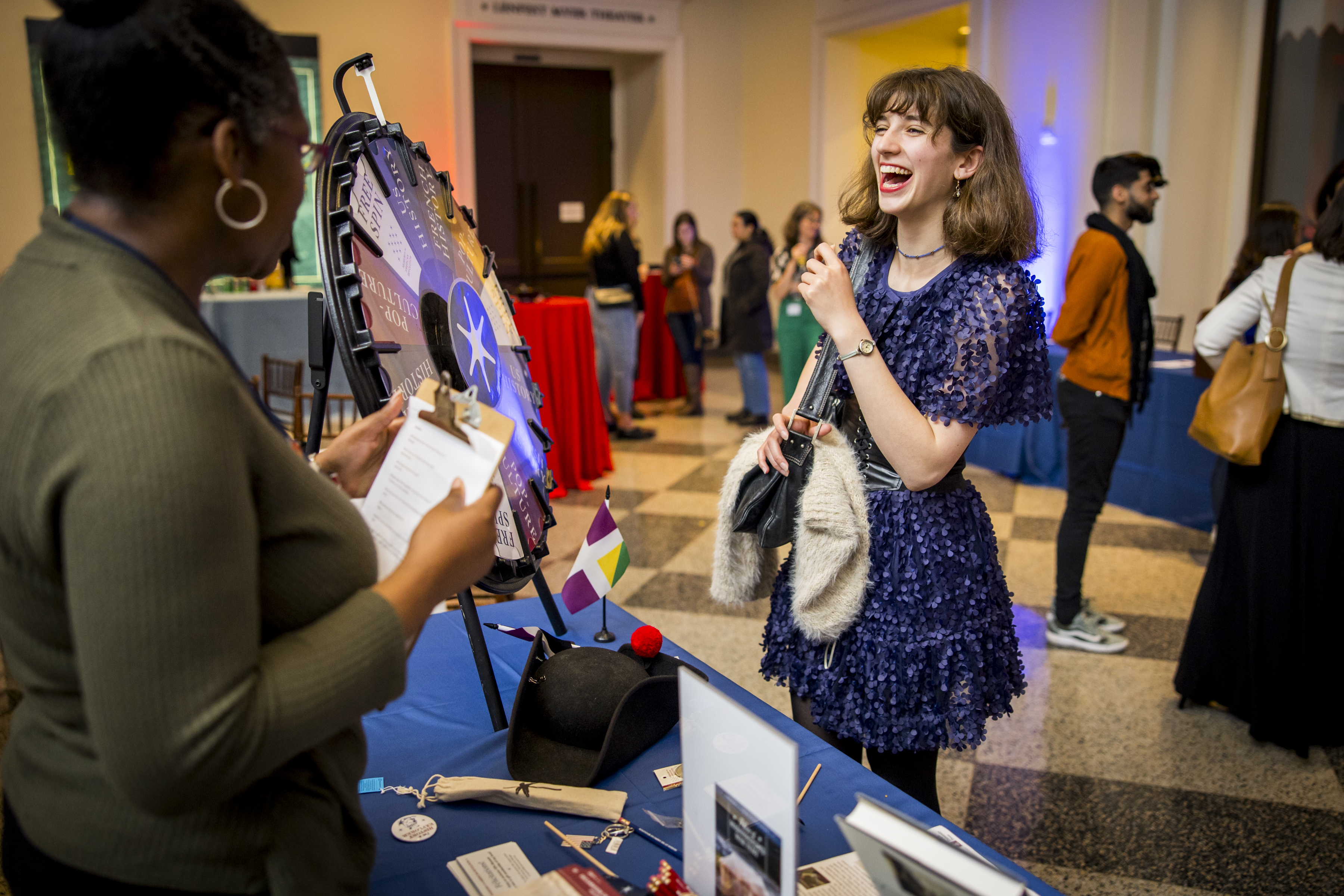 A woman in a blue dress smiles and laughs in front of a prize wheel.