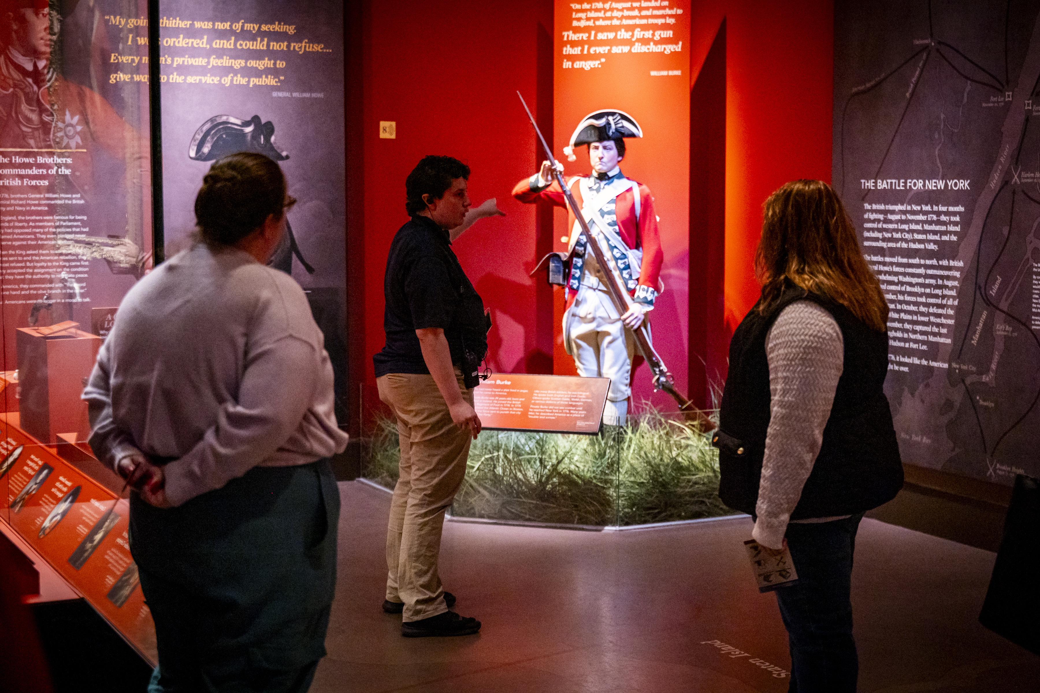 A person in a dark shirt stands in front of a group, gesturing to a life size figure in 18th century clothing