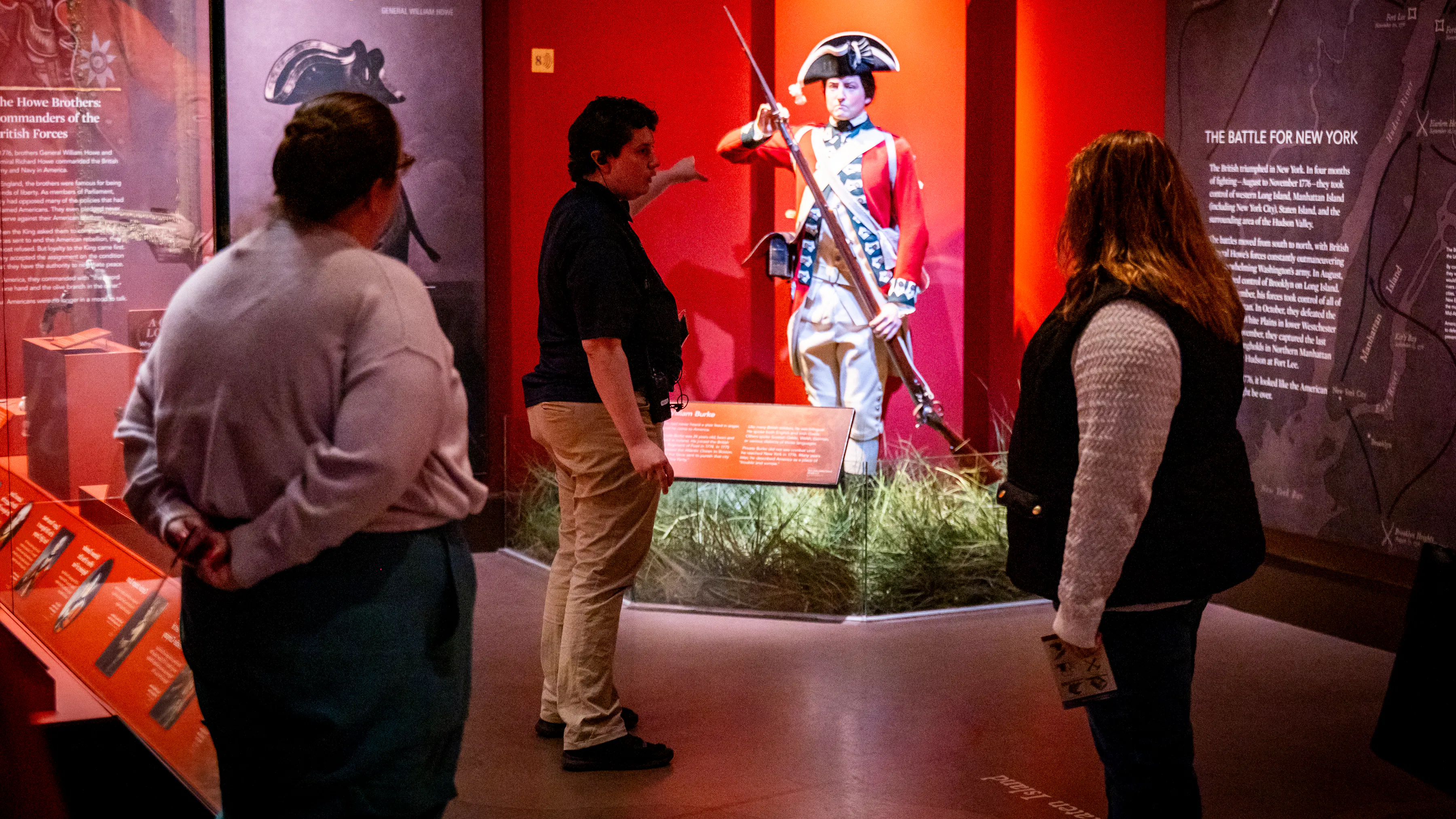 A person in a dark shirt stands in front of a group, gesturing to a life size figure in 18th century clothing