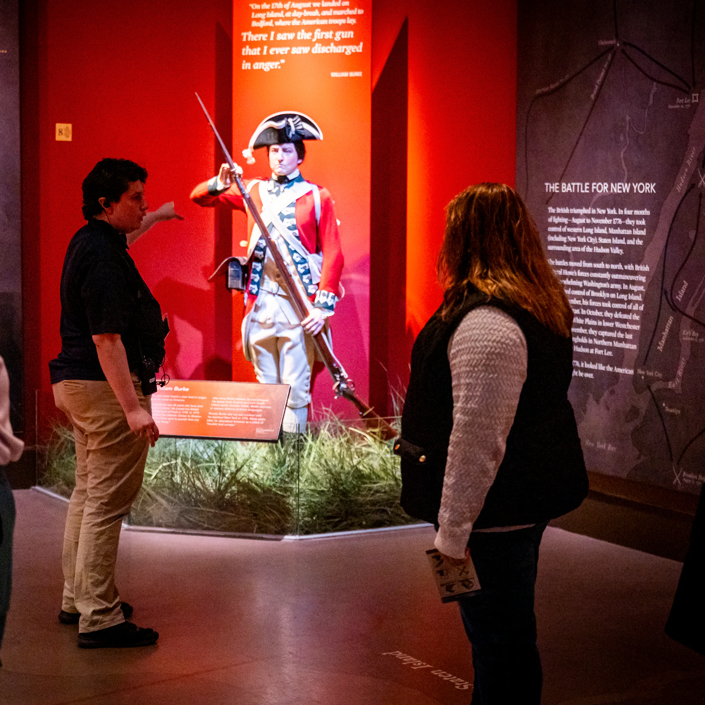A person in a dark shirt stands in front of a group, gesturing to a life size figure in 18th century clothing