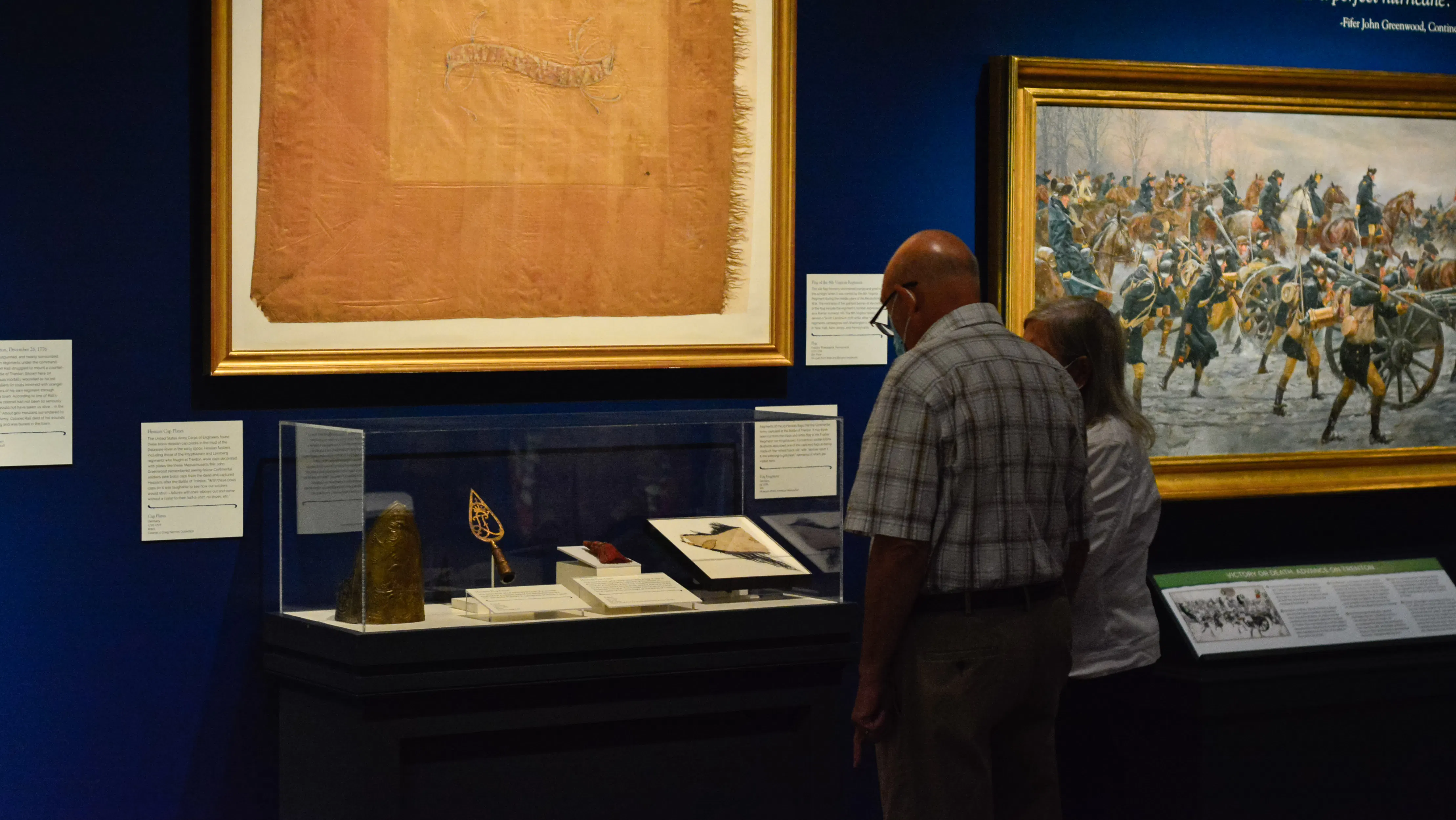 A couple looks at objects and a red and orange flag that were present at the Battle of Trenton during the Revolutionary War.