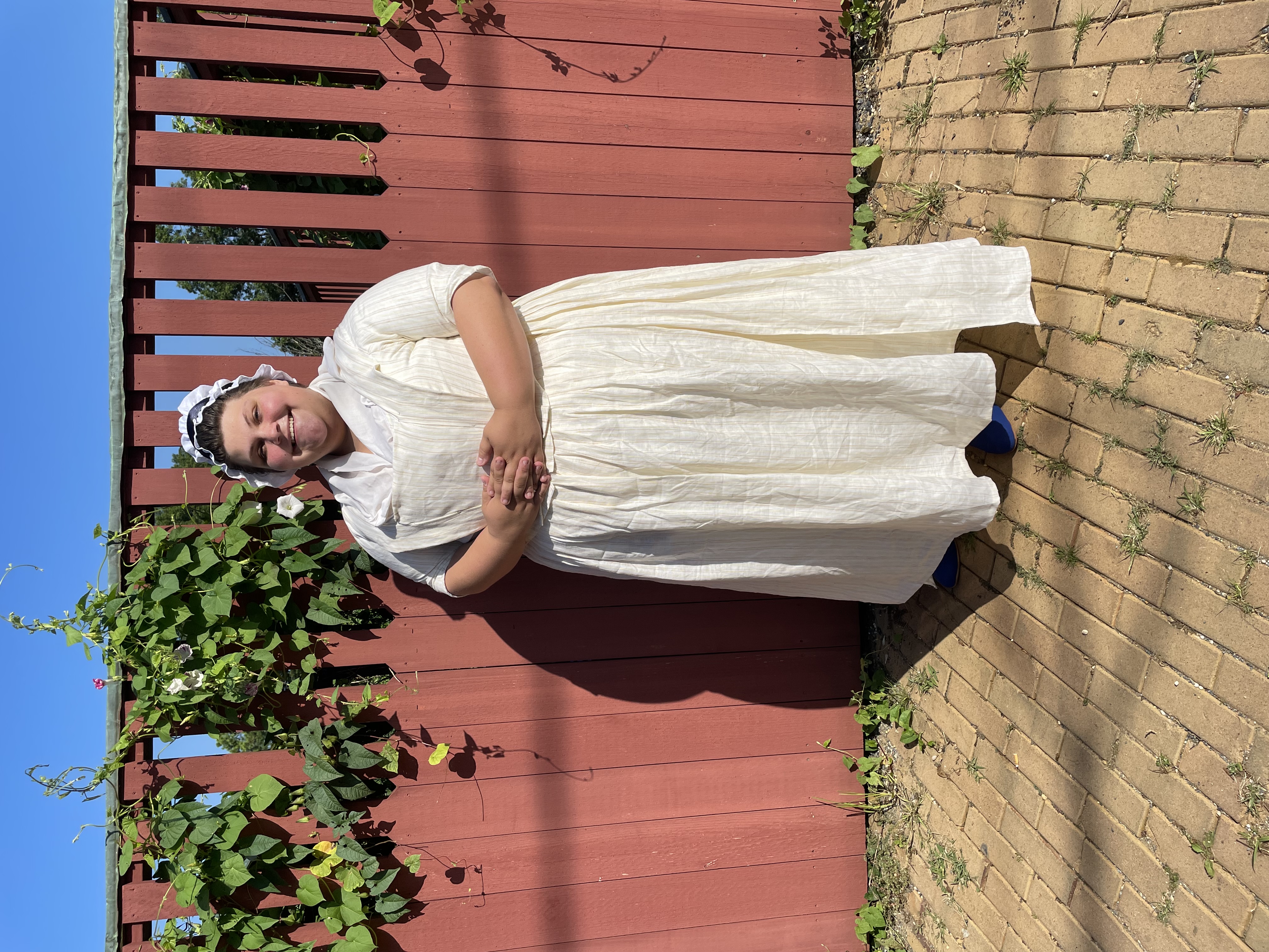 Historical interpreter Sydney Marenburg dressed in a white 18th century dress and standing in front of a red fence with green vines.