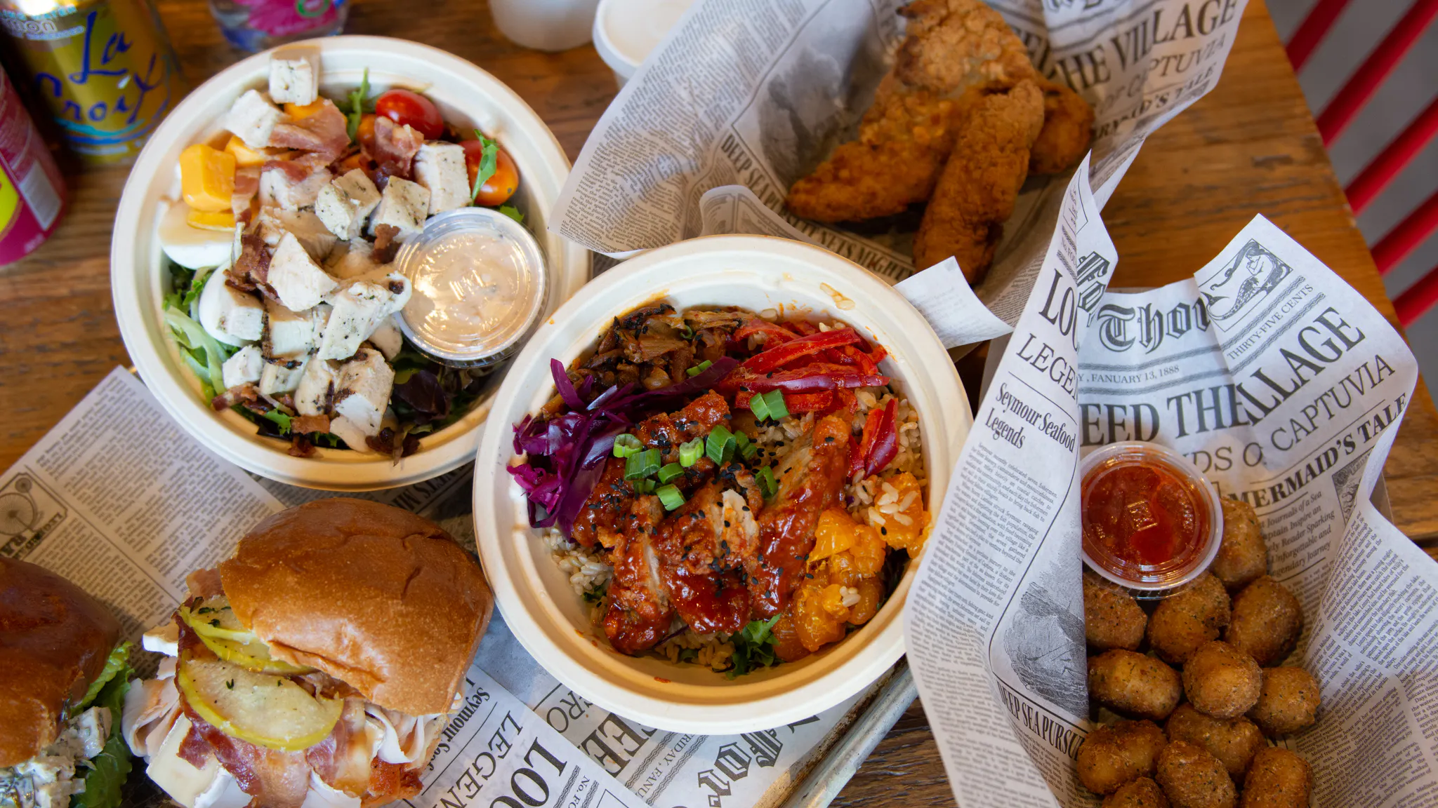Several food items arranged on a table, including salads, sandwiches and chicken fingers, several items on top of newspaper.