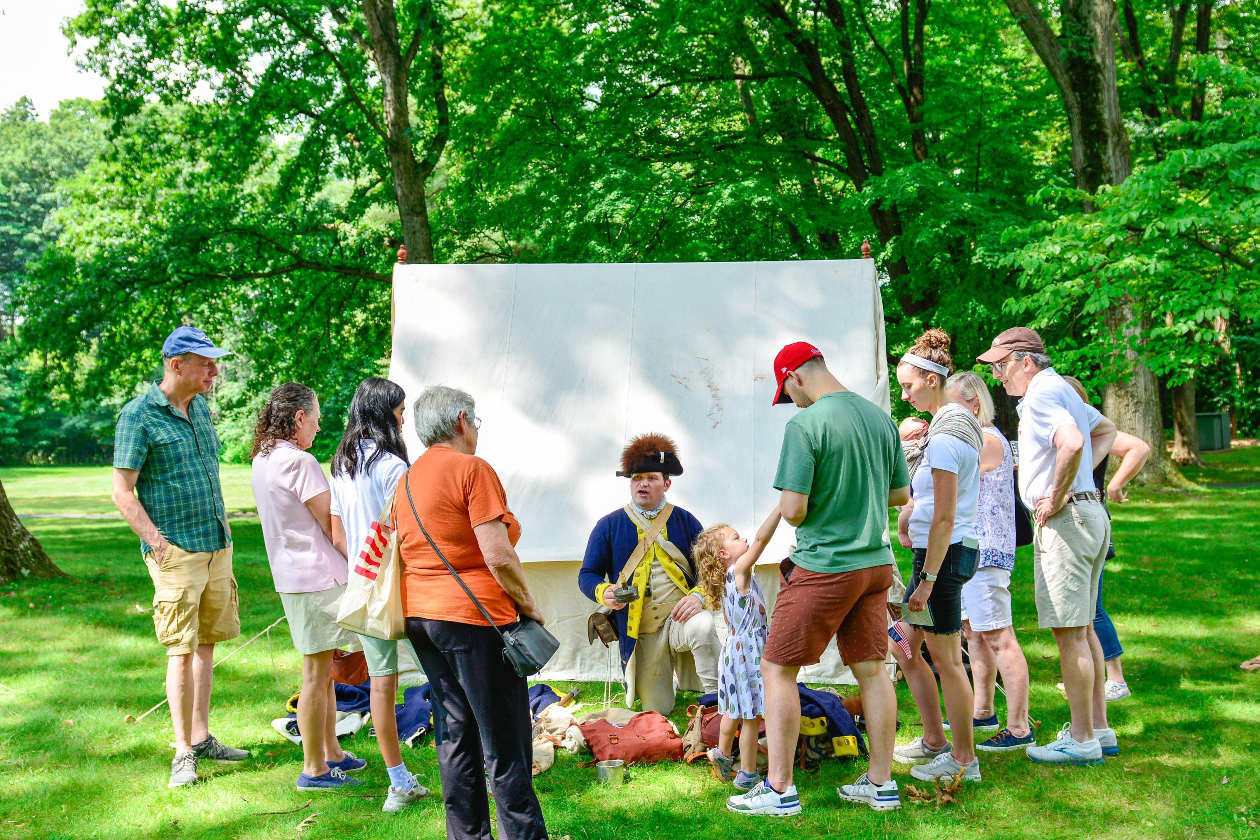 This image shows a crowd of about a dozen people in a semicircle around a costumed living historian interpreting the baggage and equipment that's part of the replica First Oval Office Project.